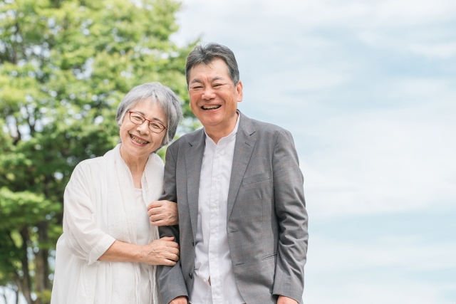 緑を背景に腕を組んで笑顔を見せる仲の良い老夫婦